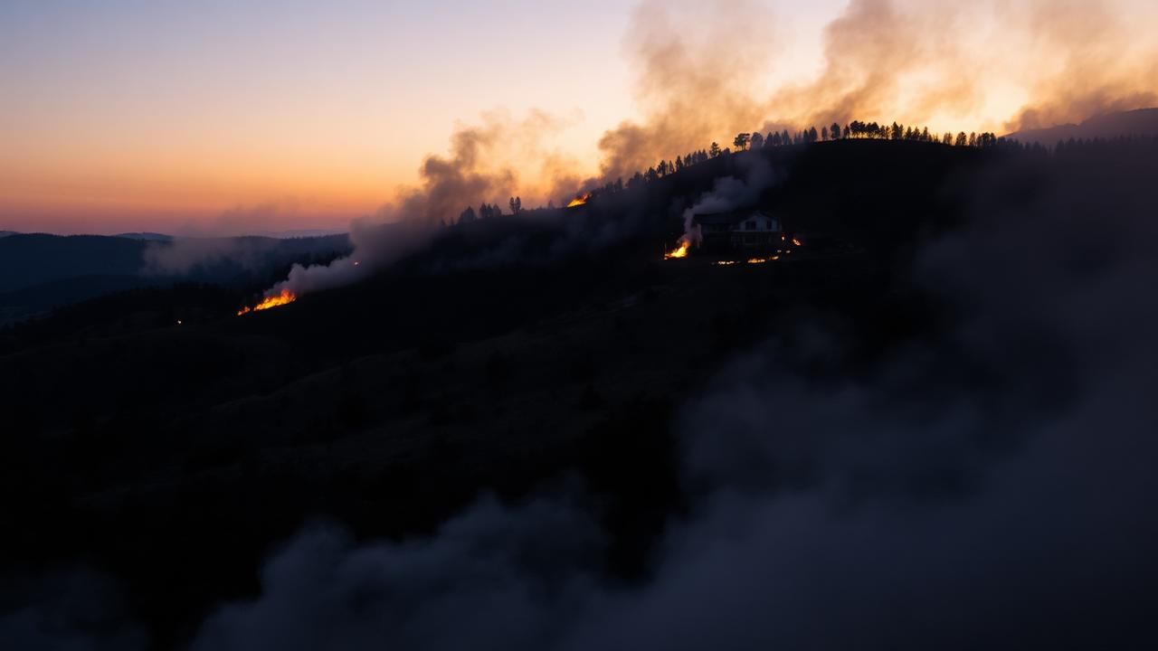 A house in the distance with smoke from a wildfire on a hillside at dusk